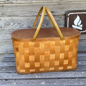 May include: A vintage wooden picnic basket with a woven design and yellow handles. The basket has a brown lid and is made of natural wood.