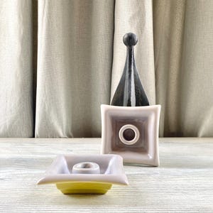 May include: Two ceramic candle holders. One is a square shape with a light purple top and a yellow base. The other is a square shape with a dark brown top and a black, pointed, decorative element.