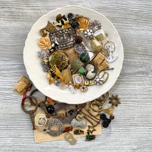 Puede incluir: Una colección de botones, alfileres y pequeños dijes metálicos vintage variados, apilados en un plato de cerámica blanca con bordes festoneados y esparcidos sobre una superficie texturizada, incluyendo formas florales y geométricas.