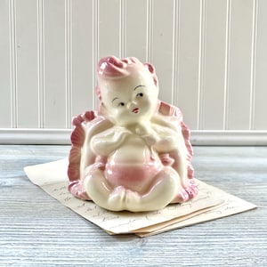 May include: A vintage ceramic baby figurine in a seated position, with a pink and cream color scheme. The baby is nestled in a pink ruffled blanket. The figurine is on a stack of aged paper.
