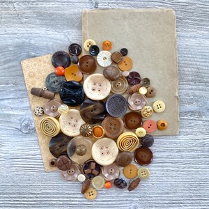 May include: A collection of vintage buttons in various sizes, shapes, and colors, including brown, beige, orange, and black. The buttons are arranged on a light brown paper and a vintage book, set against a gray wooden background.