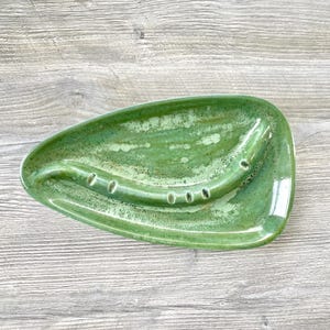May include: A handmade, ceramic soap dish in a unique, organic shape. The dish is glazed in shades of green, with a glossy finish. It features a raised, curved section with drainage holes for soap.