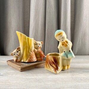 Könnte beinhalten: Vintage Keramikfiguren. Eine ist eine gelb-braune Baumstammvase mit zwei braunen Bären. Die andere ist ein kleiner Pflanztopf mit einer Jungenfigur. Der Junge trägt einen blauen Hut und ein blau-weißes Outfit. Die Figuren stehen auf einer Holzoberfläche.