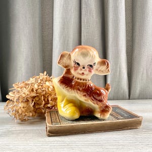 May include: A vintage ceramic puppy figurine with a brown and tan coat, sitting in a yellow boot. The puppy has large, expressive eyes and a small, brown collar. The figurine is placed on a vintage book.