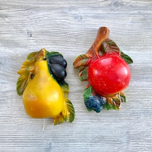 Pode incluir: Duas peças de decoração de parede vintage com frutas. Uma apresenta uma pera amarela com folhas verdes e azeitonas pretas. A outra retrata uma maçã vermelha com folhas verdes e bagas azuis. Ambas são montadas em uma superfície de madeira.