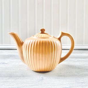 May include: A pale peach ceramic teapot with a ribbed design. The teapot has a lid and a handle.