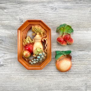 May include: A set of three decorative wall hangings. One is an octagonal frame with a relief of a pitcher, fruit, and grapes. The other two are a radish and an orange. The colors are orange, red, green, and blue.