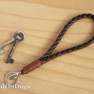 May include: A brown and black braided leather keyring with a silver key ring. The keyring is on a wooden surface.