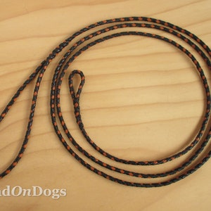 May include: A black and orange braided dog lead. The lead is made of a durable material and features a loop handle and a loop at the end for attaching to a collar. The lead is shown on a light wood surface.