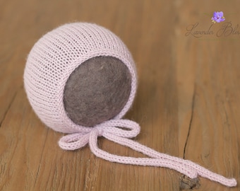 May include: A pink knitted baby bonnet with a gray felt lining and a tied bow. The bonnet is on a wooden surface.