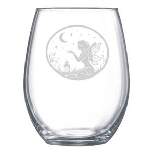 Puede incluir: Una copa de vino sin tallo transparente con un diseño esmerilado de una hada, una luna, estrellas y una linterna. El hada está arrodillada y parece estar soplando chispas. La copa tiene forma ovalada y un diseño liso y curvado.