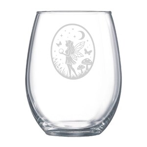 May include: A clear, stemless wine glass with an etched design. The design features a fairy holding a sphere, with butterflies, a crescent moon, stars, and mushrooms. The glass is oval-shaped and suitable for wine or other beverages.