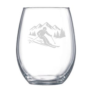 May include: A clear stemless wine glass with a skier design. The design features a skier, mountains, and pine trees. The glass is ideal for enjoying wine or other beverages.