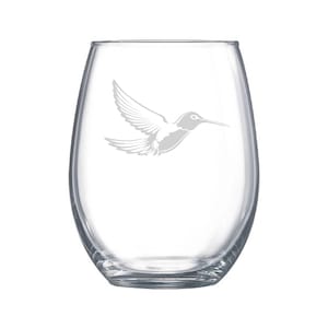 May include: A clear, stemless wine glass featuring an etched hummingbird design. The hummingbird is shown mid-flight with outstretched wings. The glass has an oval shape and a sturdy base.