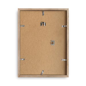 May include: A rectangular picture frame shown from the back, featuring a light brown wooden border and a tan fiberboard backing. The frame includes several silver metal swivel clips along the edges and two metal hangers for wall mounting.