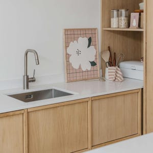 May include: A kitchen counter with a stainless steel sink and faucet, featuring a framed print of a white flower on a pink grid background. Beside the sink, a wooden shelf holds glass jars, a white bread box, and kitchen utensils in a holder.