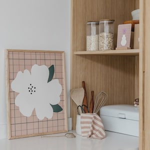 May include: A framed wall art print featuring a large white flower with dark green leaves on a pink grid background. Beside it, a wooden shelf holds two clear glass jars filled with grains, a small card with a vase illustration, and kitchen tools.