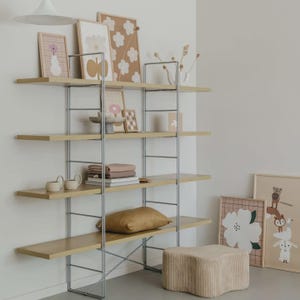 May include: A modular shelving unit with four light wood shelves supported by a silver metal frame. The shelves hold various items including framed art prints with floral motifs, a small bowl, stacked textiles, and a gold-toned throw pillow.