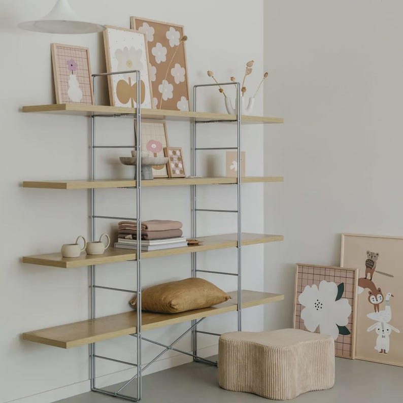 May include: A modular shelving unit with four light wood shelves supported by a silver metal frame. The shelves hold various items including framed art prints with floral motifs, a small bowl, stacked textiles, and a gold-toned throw pillow.