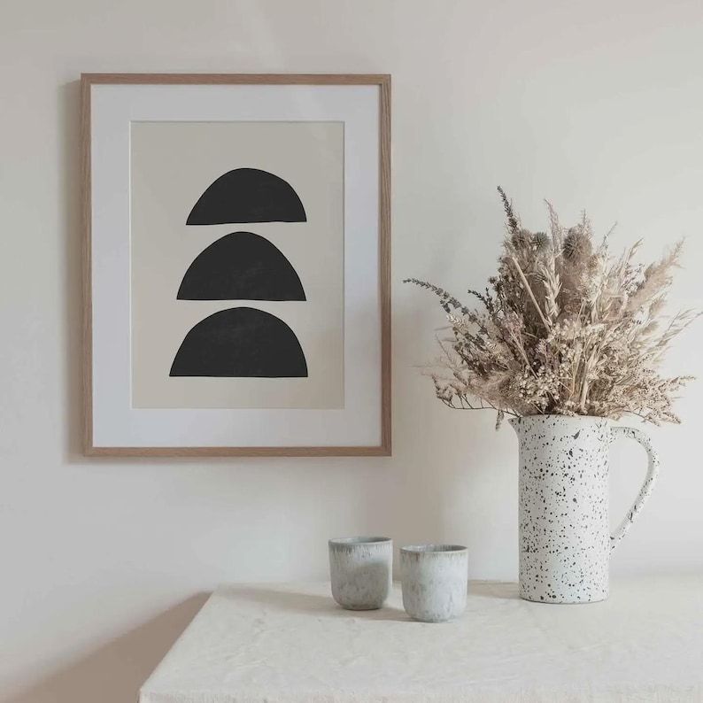 May include: A framed wall art print featuring three stacked black semi-circle shapes on a beige background. Beside the frame sits a white speckled ceramic pitcher filled with dried grasses and two small, matching speckled ceramic cups.