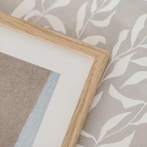 May include: A light-colored wooden picture frame with a clean mitered corner joint. The frame features a white mat board border surrounding a textured beige and light blue paper insert, showcasing a simple and neutral design aesthetic.