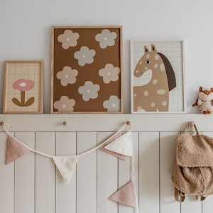 May include: Three framed wall art prints featuring a single pink flower, a brown background with light-colored flowers, and a spotted horse illustration. A fabric bunting with pink and white triangles and a small plush animal backpack hang below.