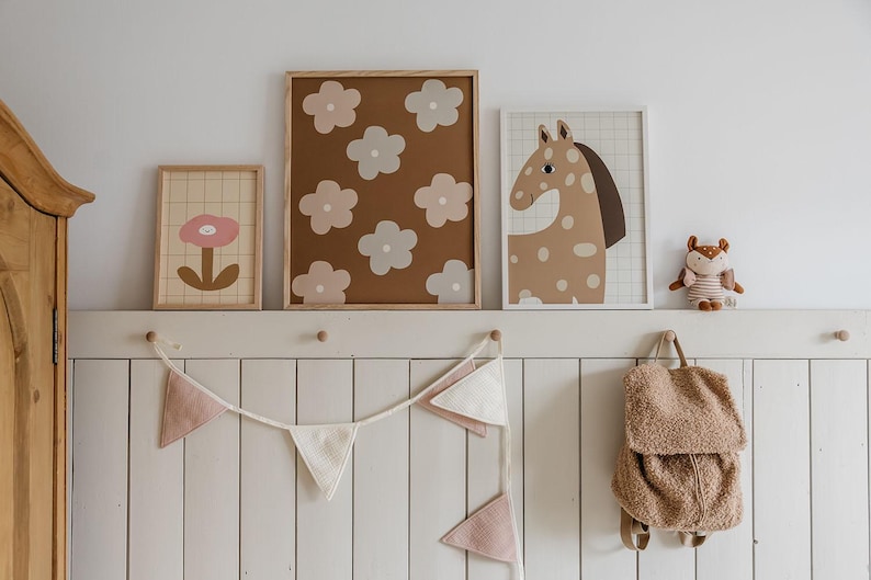 May include: Three framed wall art prints featuring a single pink flower, a brown background with light-colored flowers, and a spotted horse illustration. A fabric bunting with pink and white triangles and a small plush animal backpack hang below.