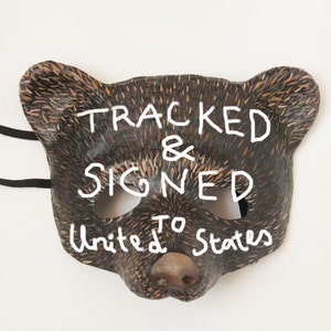 May include: A brown bear mask with white paint detailing. The mask has the words "TRACKED & SIGNED TO United States" written on it.