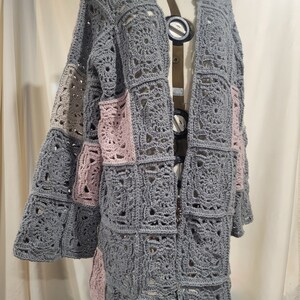 May include: A gray crocheted cardigan with pink and beige accents. The cardigan has a long, open front and a loose fit. It is made of a soft, lightweight yarn and is perfect for layering.