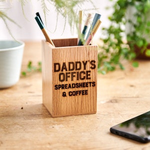 May include: A rectangular wooden pen pot with the words "DADDY'S OFFICE SPREADSHEETS & COFFEE" engraved on the front. It contains several pens and pencils. The wood has a light brown colour and a visible grain.