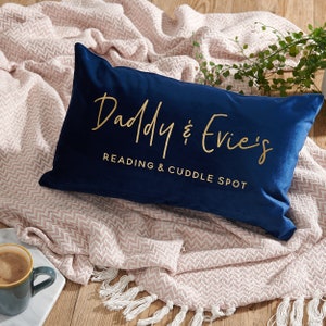May include: A navy blue velvet cushion with gold lettering that reads "Daddy & Evie's Reading & Cuddle Spot". The cushion is on a pink and white throw blanket with white fringe.