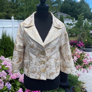 May include: A cream-colored cropped jacket with a gold floral pattern and gold buttons. The jacket is on a mannequin against a backdrop of green foliage and a white fence.