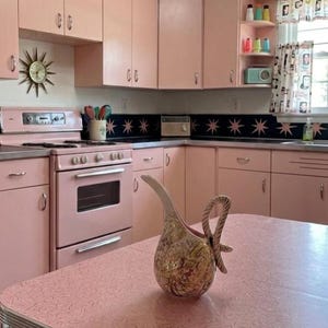 May include: A kitchen interior featuring light pink cabinets, a matching pink stove, and a pink boomerang-patterned table. A ceramic pitcher with a textured, multi-colored abstract pattern sits on the table. Two starburst wall clocks are mounted on the wall.