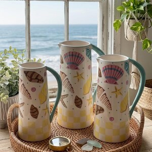May include: Three ceramic pitchers with sea-themed designs, including seashells and starfish, sit on a woven tray. The pitchers have light blue handles and are decorated with a yellow and white checkered pattern at the base. The background shows a window with a view of the ocean.