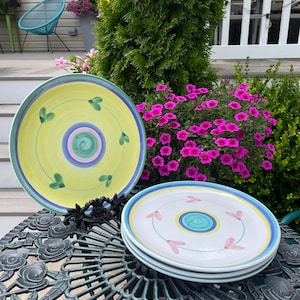 May include: A stack of white ceramic plates with a blue, yellow, and pink floral design. One plate is yellow with a green leaf design. The plates are on a decorative metal table, with flowers and greenery in the background.