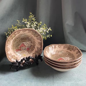 May include: A set of vintage ceramic bowls with a scenic design. The bowls feature a cream-colored interior with a brown border and a detailed illustration of a red covered bridge. One bowl is displayed on a black decorative stand.