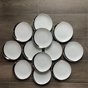 May include: A set of ten white ceramic plates with a black and silver trim. The plates are arranged in a circle on a wooden surface.
