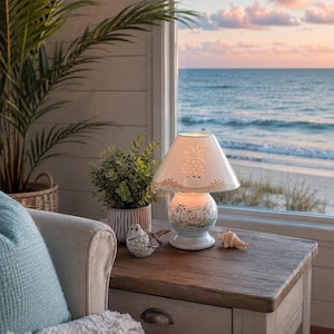 May include: A decorative table lamp with a pineapple design on the shade and a beach scene on the base. The lamp sits on a wooden table next to a small bird figurine and a seashell. The background shows a beach and ocean view.
