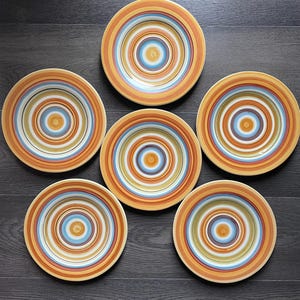 May include: Six ceramic plates with concentric circles in shades of orange, yellow, blue, and white. The plates are arranged on a dark wood surface, showcasing a vibrant and colorful design. Ideal for serving meals.