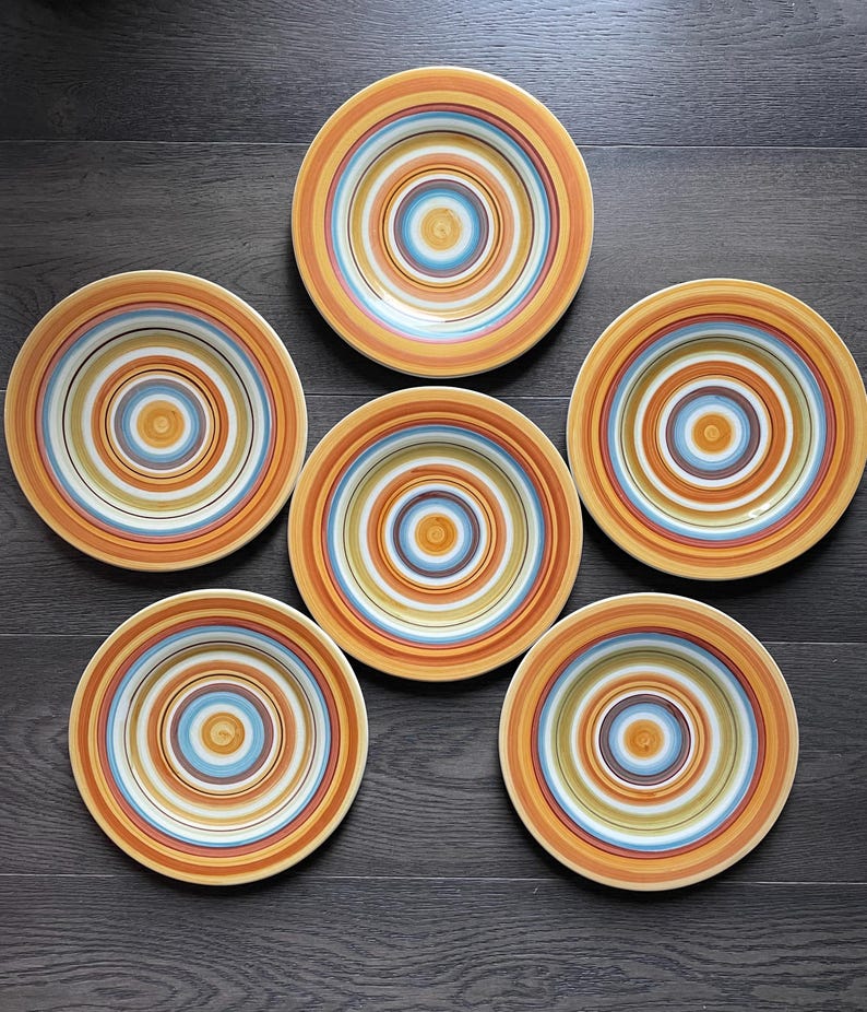 May include: Six ceramic plates with concentric circles in shades of orange, yellow, blue, and white. The plates are arranged on a dark wood surface, showcasing a vibrant and colorful design. Ideal for serving meals.
