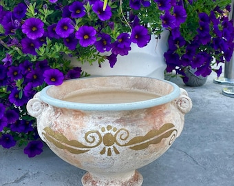 May include: A decorative, aged-looking terracotta planter with a light blue rim and gold leaf design. The planter is filled with a light brown substance and sits in front of a white planter overflowing with vibrant purple flowers.