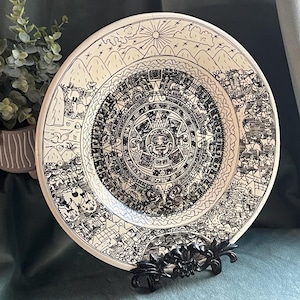 May include: A decorative ceramic plate featuring a detailed black and white illustration of the Aztec calendar. The plate is round, with a cream-colored background and a black metal stand. The design includes mountains, sun, and intricate patterns.