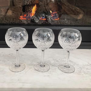 May include: Three clear crystal wine glasses with etched patterns. The glasses have a round bowl shape, a long stem, and a circular base. The etched design features a symmetrical pattern. The glasses are displayed on a white surface with a fireplace in the background.