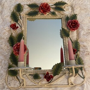 May include: White metal wall mirror with a rope-like frame and floral accents. The mirror has two candle holders with pink candles. The floral accents are made of metal and painted in shades of green and red.