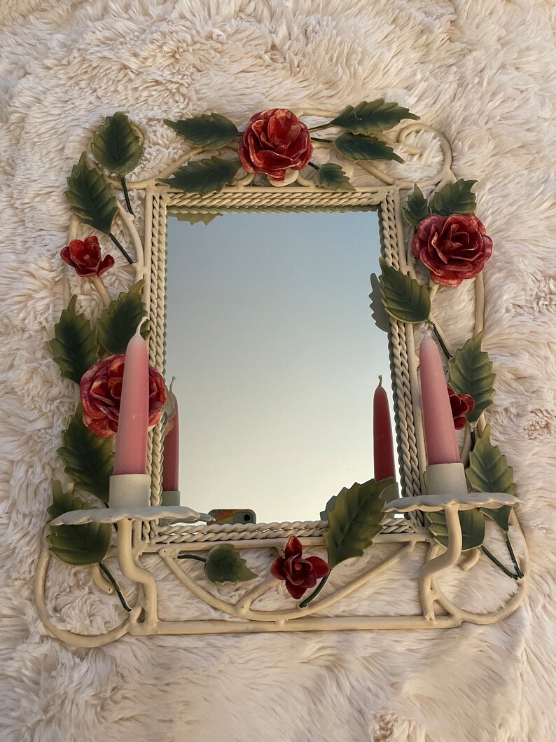May include: White metal wall mirror with a rope-like frame and floral accents. The mirror has two candle holders with pink candles. The floral accents are made of metal and painted in shades of green and red.