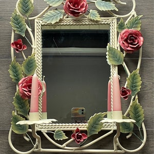 May include: White metal mirror with a floral design. The mirror has a white rope border and features pink roses and green leaves. Two pink candles are on either side of the mirror.