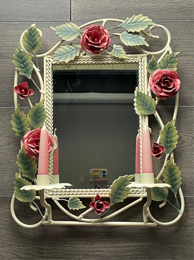 May include: White metal mirror with a floral design. The mirror has a white rope border and features pink roses and green leaves. Two pink candles are on either side of the mirror.