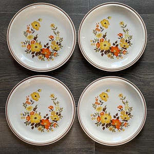 May include: Set of four white ceramic dinner plates with a brown rim and a floral design featuring yellow, orange, and brown flowers.