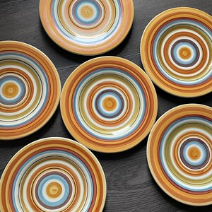 May include: Six ceramic plates with concentric circles in shades of orange, brown, blue, and white. The plates have a rustic, handmade appearance, perfect for serving meals. The plates are arranged on a dark wooden surface.