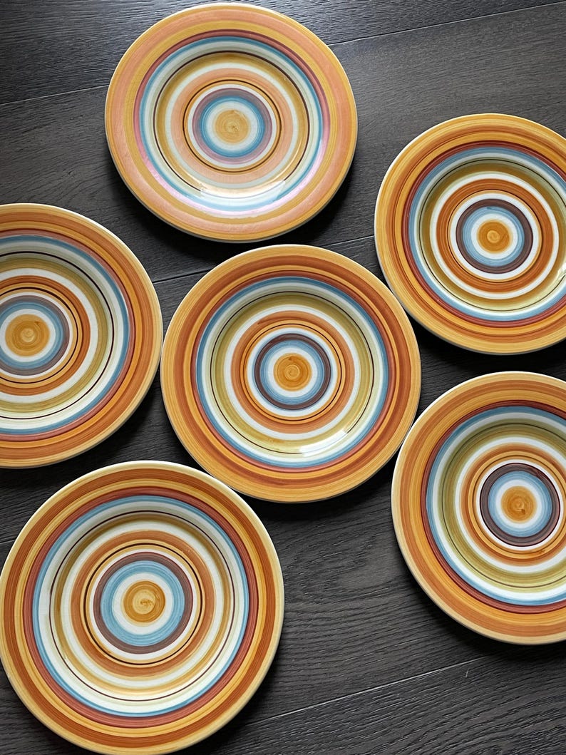 May include: Six ceramic plates with concentric circles in shades of orange, brown, blue, and white. The plates have a rustic, handmade appearance, perfect for serving meals. The plates are arranged on a dark wooden surface.